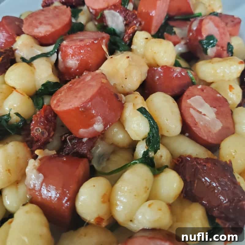 A close-up of Smoked Sausage Gnocchi with sun-dried tomatoes and spinach on a white plate, garnished with fresh herbs and Parmesan, ready to eat.