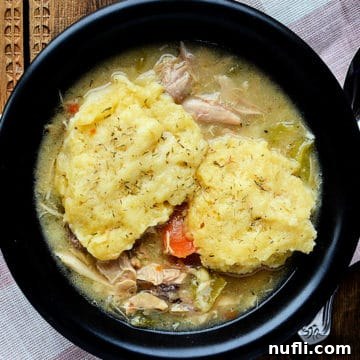 Crockpot chicken and dumplings in a dark bowl next to a spoon, garnished with fresh herbs