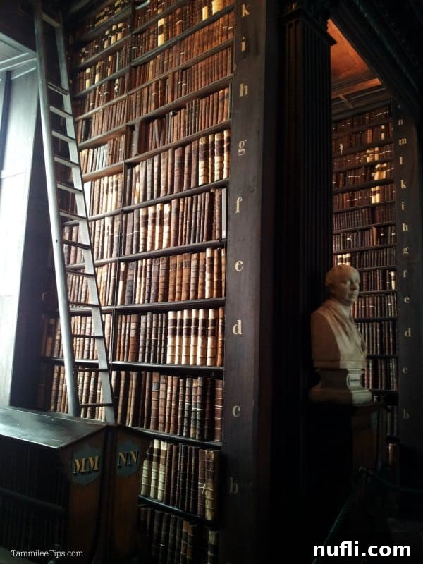 Interior view of the Long Room at Trinity College Library, showcasing towering shelves of alphabetized books, a vintage wooden rolling ladder, and a classical bust at the end of a book-lined row.