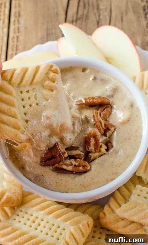 A crispy cookie being dipped into the rich, warm pecan praline dip, with whole pecans visible on the surface of the creamy dessert.