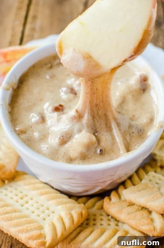 A vibrant red apple slice generously coated in warm praline dip, surrounded by a variety of cookies and a white bowl of dip.
