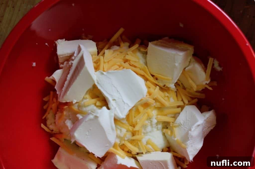 Cream cheese and shredded cheddar cheese mixed into mashed potatoes in a red mixing bowl, ready for topping.
