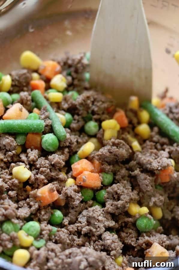 Cooked ground beef and mixed vegetables combined with beef gravy in a glass bowl, stirred with a wooden spoon.