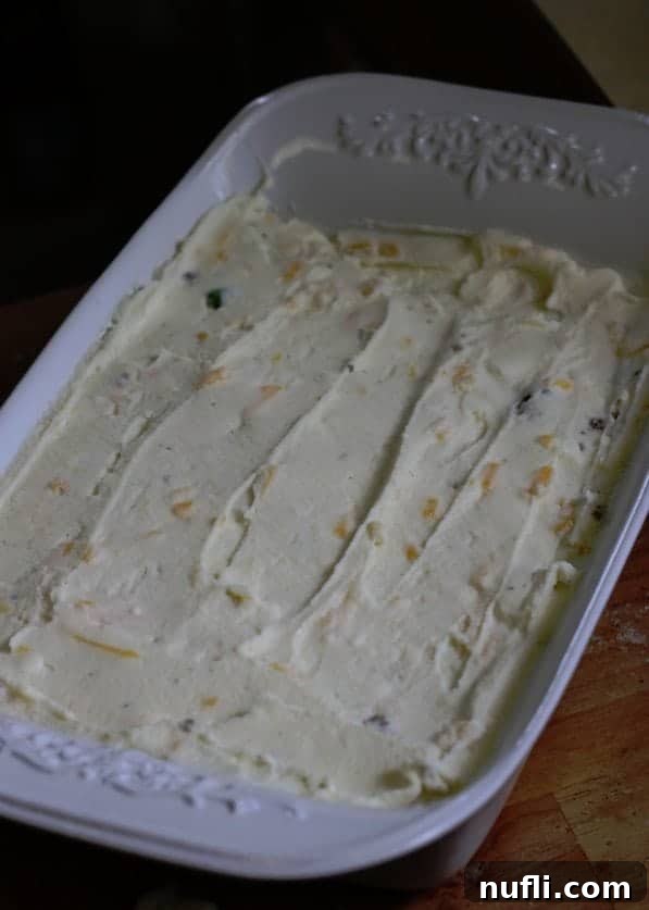 Creamy mashed potatoes with cheese being spread over the ground beef layer in a white baking dish.