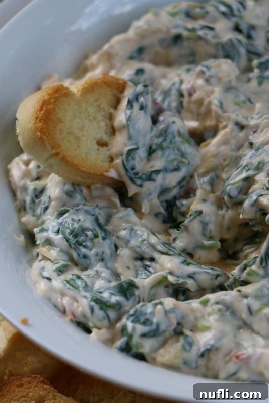 Toasted bread pieces being dipped into a large bowl of Creamy Spinach Artichoke Dip, presented on a wooden board.