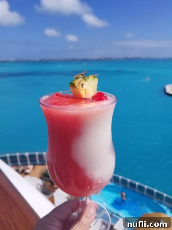 A hand holding a Miami Vice Cocktail in a hurricane glass above a cruise ship railing, with the vast, blue ocean sparkling in the background.