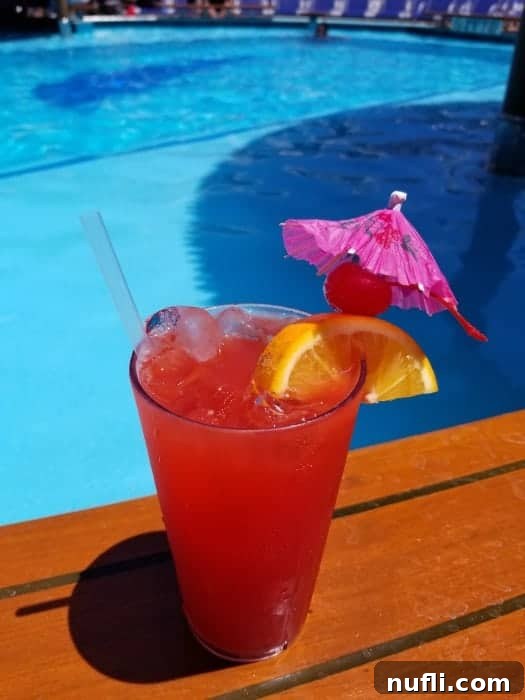 A Carnival Fun Ship Cocktail with an orange wedge, cherry, and umbrella garnish, enjoyed by a cruise ship pool