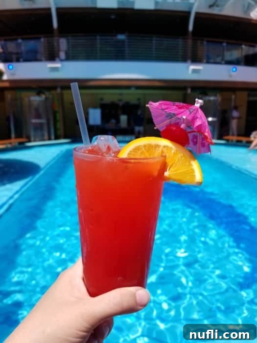 A hand holding a red tropical drink, garnished with an orange slice, cherry, and a pink umbrella, by a pool