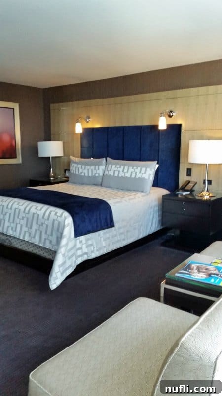 Aria Hotel Room with king bed, blue headboard, night stands with lamps and a chair and table 