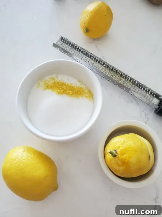 Fresh lemon zest and sugar in a white bowl, with a microplane zester and whole lemons in the background, highlighting key ingredients for lemon sugar cookies.
