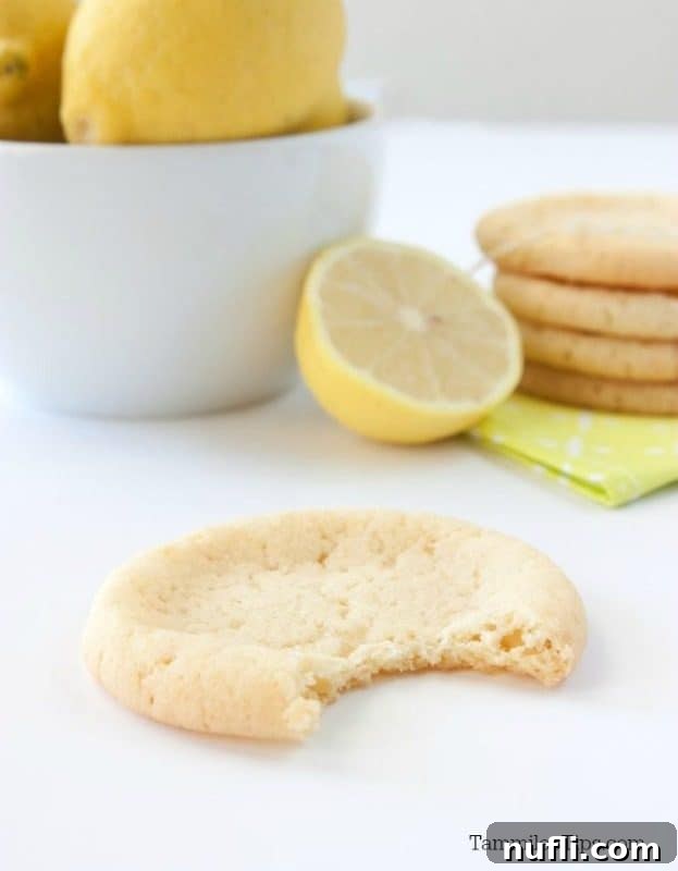 A half-eaten Lemon Sugar Cookie next to a bowl of fresh lemons and a stack of baked cookies, showcasing the deliciousness.