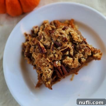 Close-up of a perfectly baked square pumpkin pie bar on a white plate, garnished with a small pumpkin and autumn leaves.