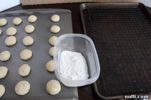 Freshly baked Snowball cookies on a baking sheet, with a bowl of powdered sugar and a wire cooling rack nearby
