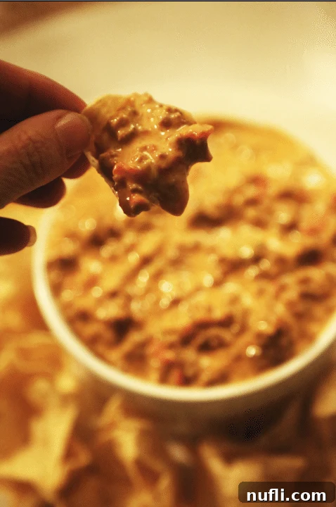 Easy Crock Pot Chili Cheese Dip 2 Hand holding a chip over a bowl of Crockpot Chili Cheese Dip, ready to be enjoyed.