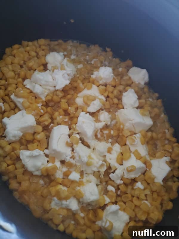 Uncooked crockpot creamed corn in a black slow cooker, showing chunks of cream cheese spread over the frozen corn kernels before cooking.
