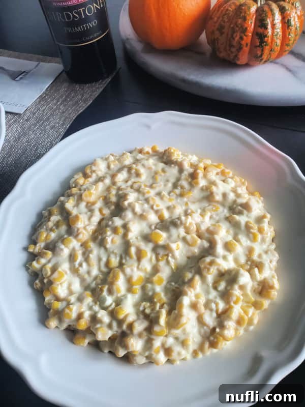 Creamy Crockpot Creamed Corn in a white serving bowl with autumn decorations like pumpkins and a bottle of wine in the background.