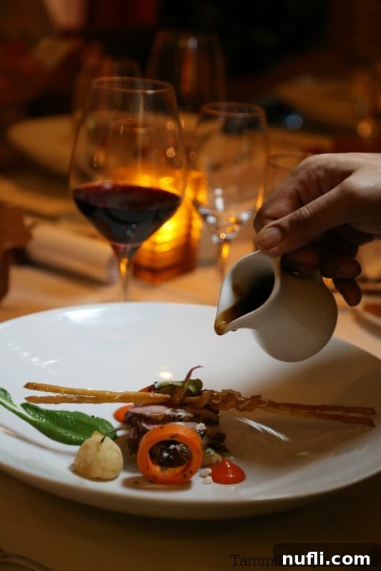 A chef's hand skillfully pours a rich sauce over a beautifully arranged gourmet dish of meat and vibrant vegetables.