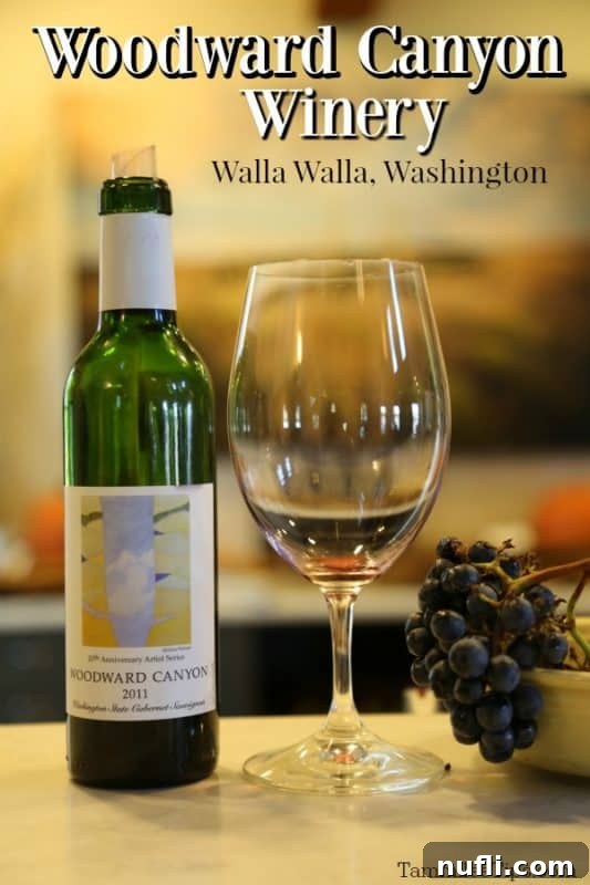 A bottle of Woodward Canyon wine and a wine glass elegantly displayed, with the winery's historic building visible in the soft focus background, embodying the essence of Walla Walla wine country.