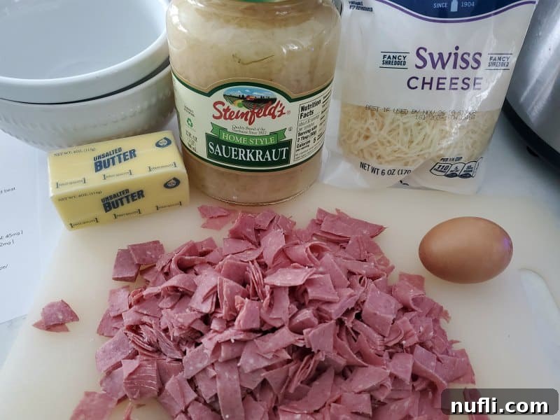 Easy Crock Pot Reuben Dip 3 Fresh ingredients for Reuben dip: a block of Swiss cheese, a package of corned beef lunchmeat, a jar of sauerkraut, a stick of butter, and a single egg, all laid out on a wooden cutting board.