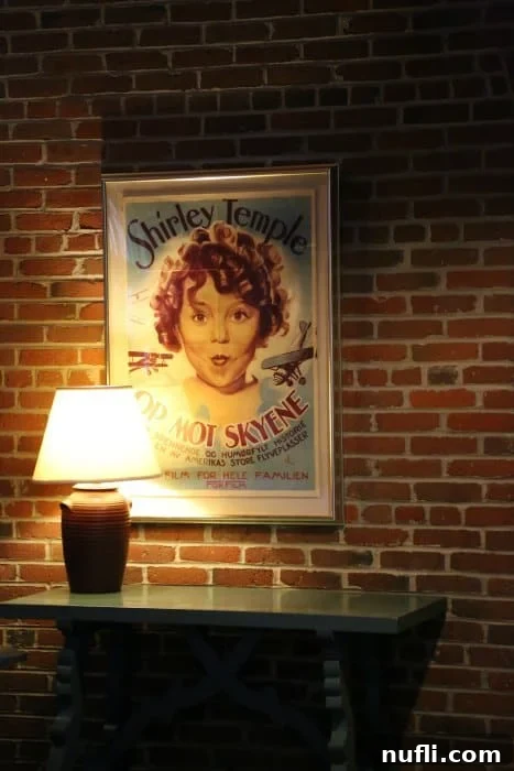 A vintage Shirley Temple movie poster, vibrant with classic Hollywood charm, displayed elegantly above a side table against a warm brick wall, showcasing a piece of cinematic history.