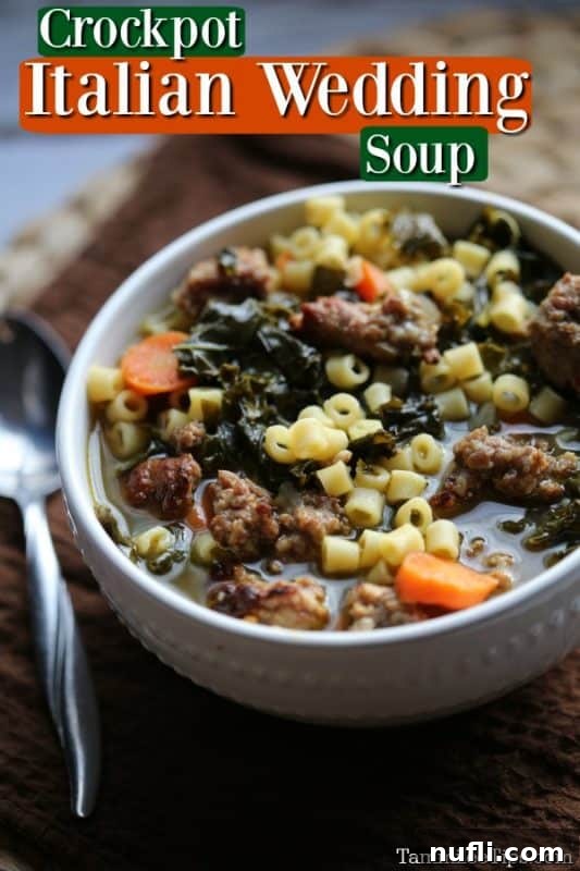 A bowl of delicious Crockpot Italian Wedding Soup, with visible Italian sausage, kale, and carrots, next to a spoon on a white surface.