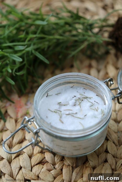 Homemade rosemary bath salts in a clear glass container, with fresh rosemary sprigs beside it, highlighting natural ingredients.