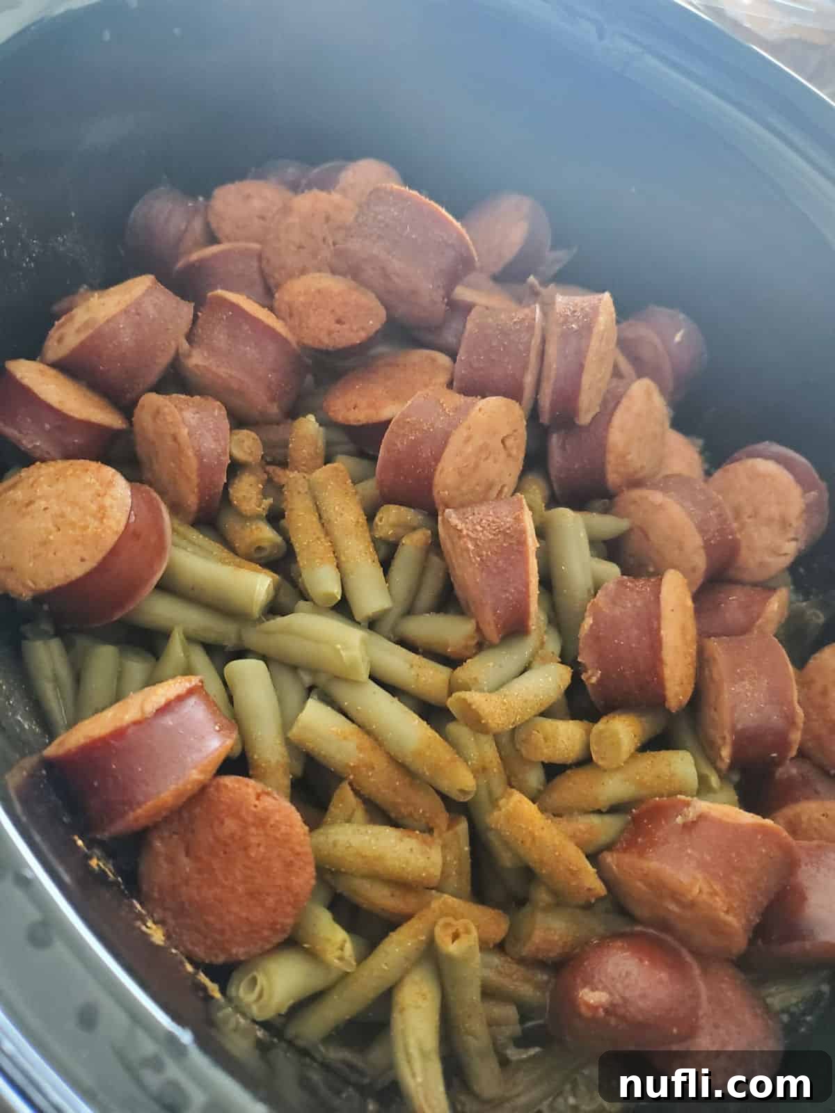 Cooked kielbasa pieces with tender green beans and potatoes in a slow cooker, steam rising, ready to serve.