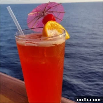 red cocktail with an orange wheel, cherry and pink umbrella with the ocean behind it