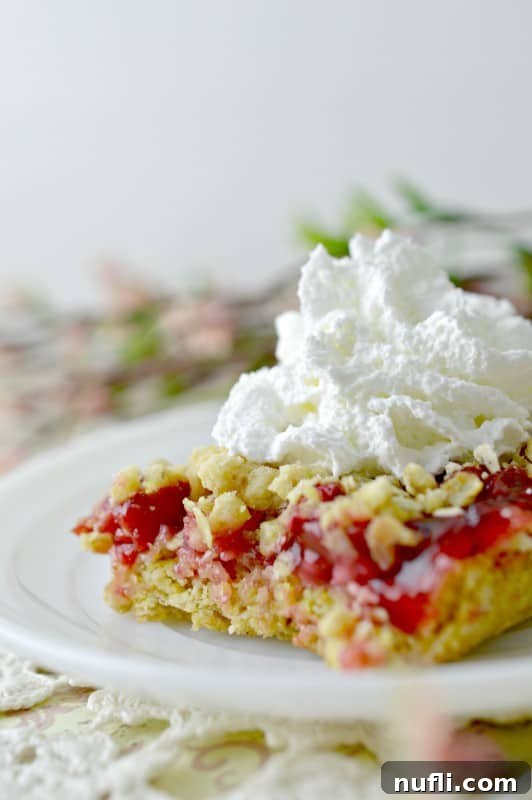 A cherry cake bar topped with a dollop of cool whip on a white plate, with a silver spoon.