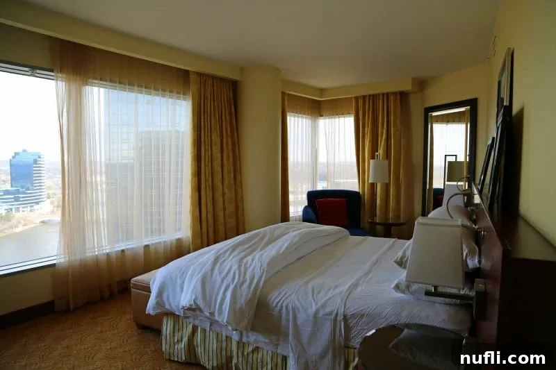 An elegant hotel room at JW Marriott Grand Rapids featuring a king-size bed, comfortable armchair, and large windows overlooking the river.