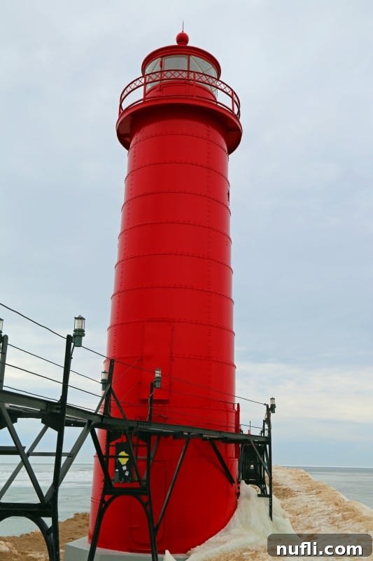 Bright red Grand Haven Lighthouse encased in a fortress of ice, with the frozen pier extending from the foreground under a clear winter sky.