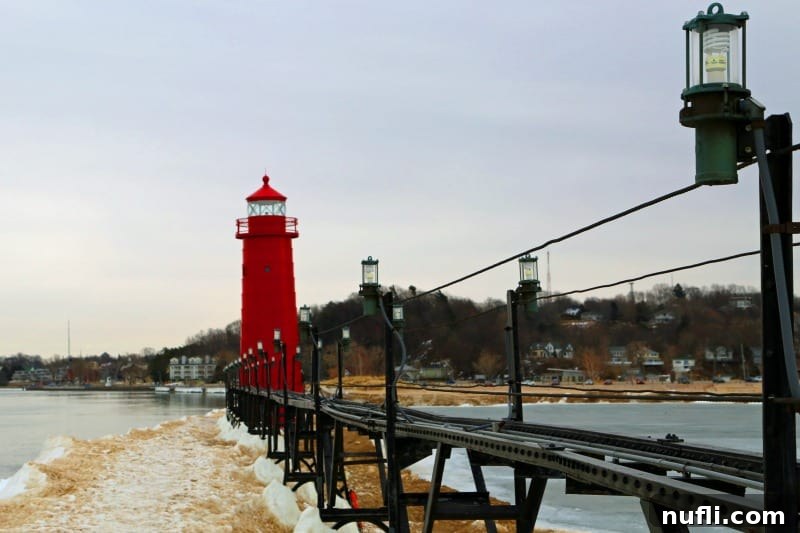 The Grand Haven Lighthouse stands resolute at the end of an ice-covered walkway, surrounded by a dramatic, frozen winter landscape.