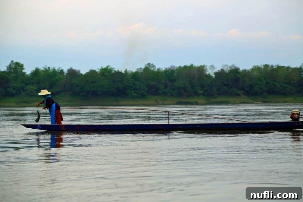 Mighty Mekong Voyage: Thailand to Laos 8 A Thai fisherman stands poised at the back of his boat on the Mekong River, preparing his fishing lines amidst the tranquil morning mist.