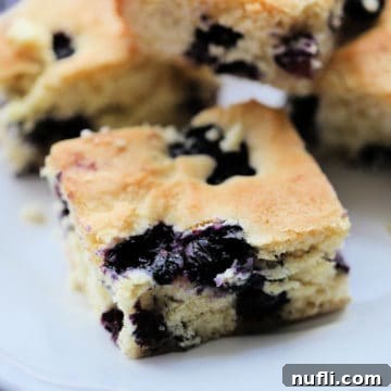 Ultimate Blueberry Cake 4 A close-up image of a square blueberry cake on a white plate, highlighting its texture and juicy fruit.