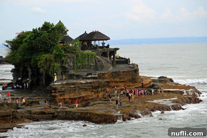 Tanah Lot Temple standing majestically on a rock formation surrounded by ocean waves, with people exploring the base