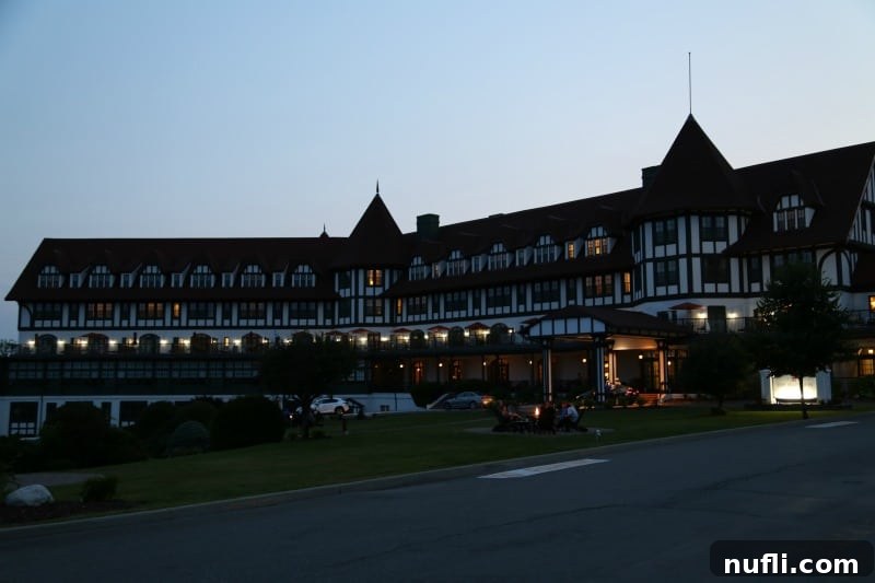 The Algonquin Resort's exterior at dusk, with soft lighting illuminating the classic Tudor architecture and a warm glow from outdoor fire pits where guests can relax.